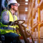 Show a professional forklift operator wearing a hard hat, reflective vest, and safety goggles, operating a forklift in a well-lit and organized warehouse setting. This image portrays the importance of safety and professionalism in forklift operations.